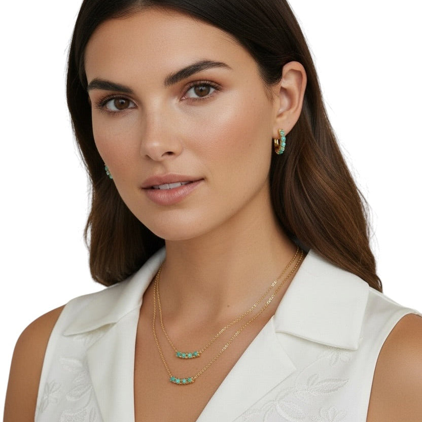 A woman with dark brown hair is wearing a white sleeveless embroidered top and layered gold necklaces with turquoise accents, along with matching turquoise hoop earrings, against a plain white background.