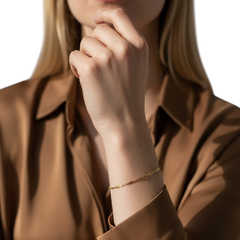A close-up of a woman wearing a delicate gold chain bracelet on her wrist, paired with a brown satin button-up shirt against a white background.