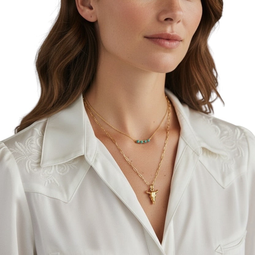 A close-up of a woman wearing layered gold necklaces, including a delicate chain with a small cross pendant and a second chain with turquoise stones, paired with a white embroidered collared shirt against a white background.