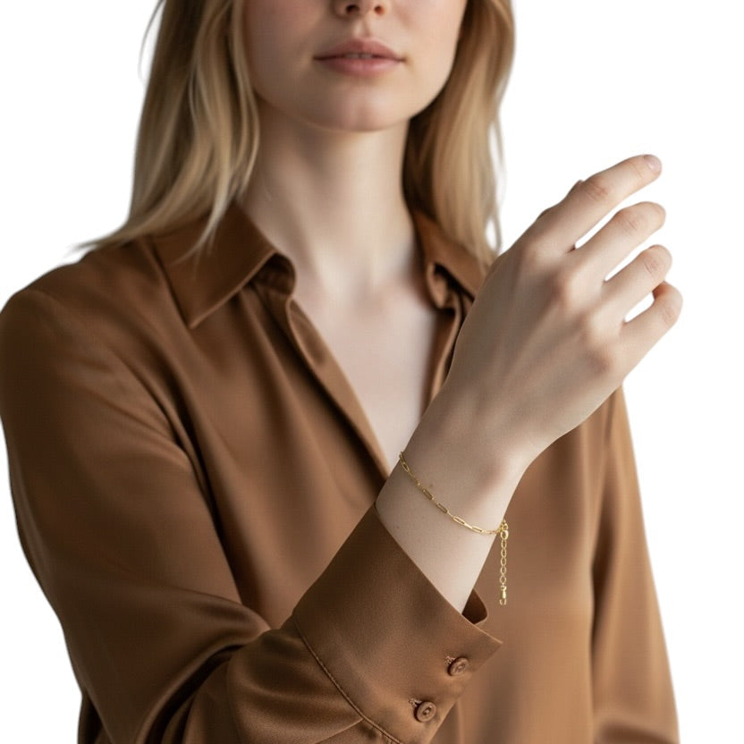 A blonde woman wearing a brown button-up shirt and a delicate gold chain bracelet, photographed against a clean white background.