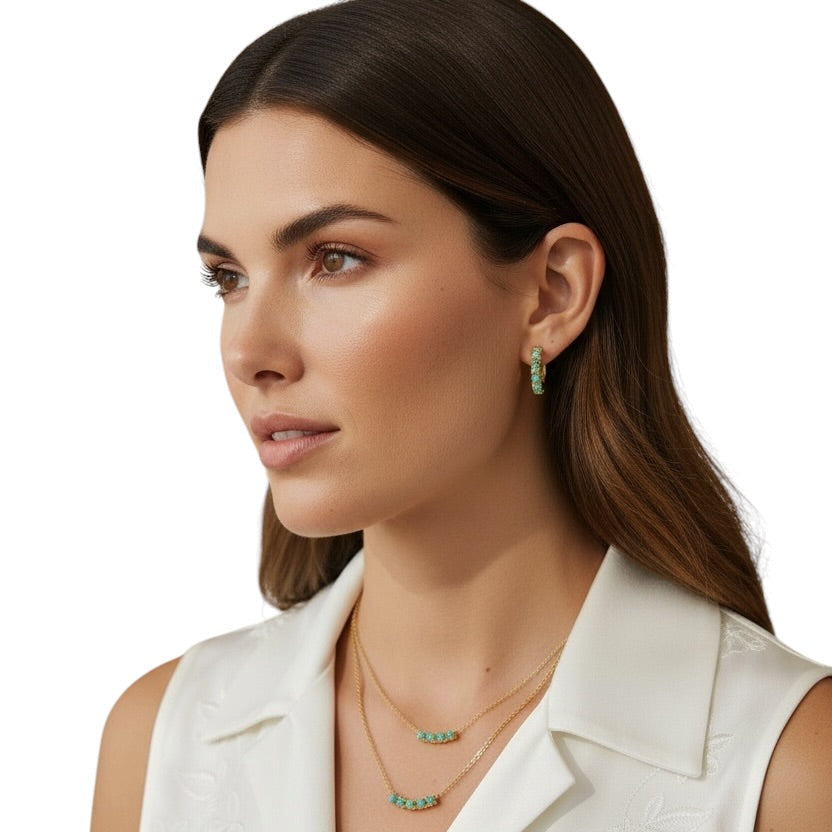A woman with long dark brown hair is shown in a close-up profile wearing a white sleeveless collared top and layered gold necklaces with small turquoise-green bead accents, along with matching drop earrings, against a plain white background.