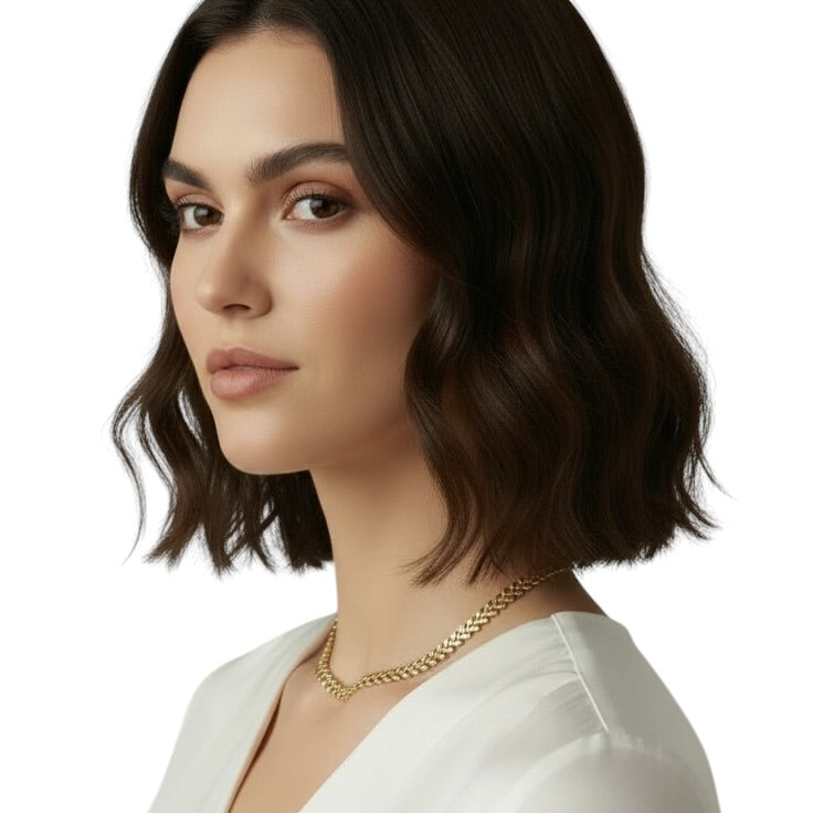 A woman with dark brown shoulder-length wavy hair is shown in a close-up portrait wearing a delicate gold chain necklace and a white top against a plain white background.